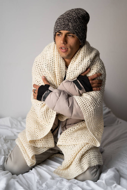 Young man warmly dressed is feeling cold, covered with warm blanket, frozen sitting on the bed at cold home. Have problem with health, central heating concept - Photo, Image