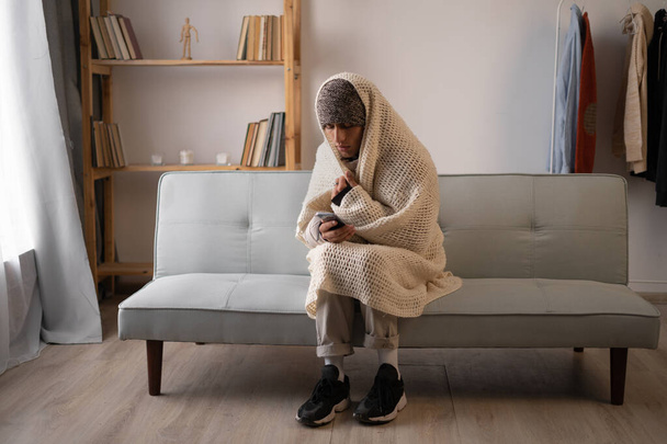 Unwell man renter in blanket sit in cold living room use mobile phone.suffer from lack of heat and power. Unhealthy young man struggle from chill freeze at home. No heating concept. - Photo, Image