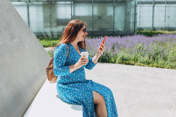 Портрет красивої молодої рудої жінки, яка посміхається, сидячи надворі на сучасній лавці з мобільним телефоном. Дівчинка п'є чашку кави. Щасливі жінки, які користуються телефоном у європейському місті.. - Фото, зображення