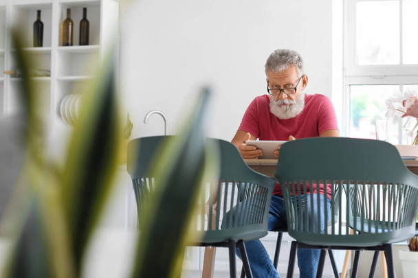 Senior man met tablet computer aan tafel in de keuken - Foto, afbeelding