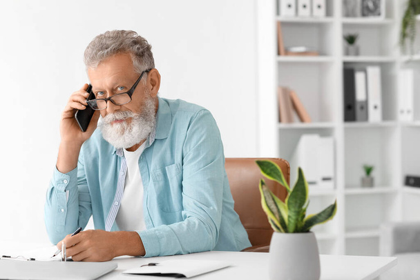 Idősebb férfi beszél mobiltelefonnal és írás az asztalnál az irodában - Fotó, kép