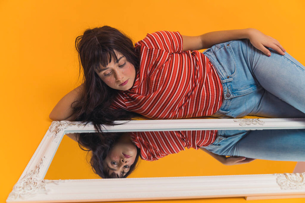 dark-haired Caucasian girl in casual clothes laying down on the floor and looking at the mirror confidently, cowboy shot studio shot orange background. High quality photo - Photo, Image