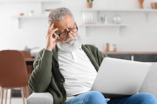 Задумчивый взрослый мужчина с ноутбуком, сидящий на диване на кухне - Фото, изображение
