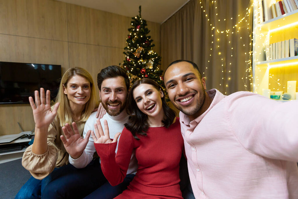 Diverse friends celebrating christmas and new year together, group of people having dinner at home sitting on sofa near Christmas tree, taking selfie photo and talking with friends on video call using - Fotografie, Obrázek