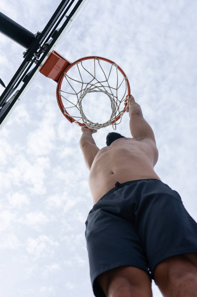 Shirtless man hanging from a basketball hoop to score on an urban court in the morning - Fotoğraf, Görsel