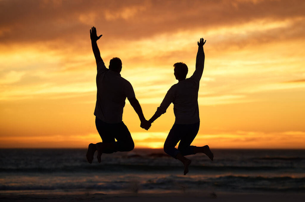 Couple, sunset and jump at beach while holding hands for happiness, bonding or romance on vacation. People, love and happy by sea, ocean or dusk in evening, sunshine or silhouette on holiday together. - 写真・画像