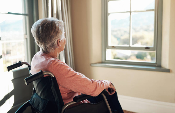 Quem me dera ter a minha vida de volta. uma mulher idosa em uma cadeira de rodas olhando pensativamente para fora de uma janela em casa - Foto, Imagem