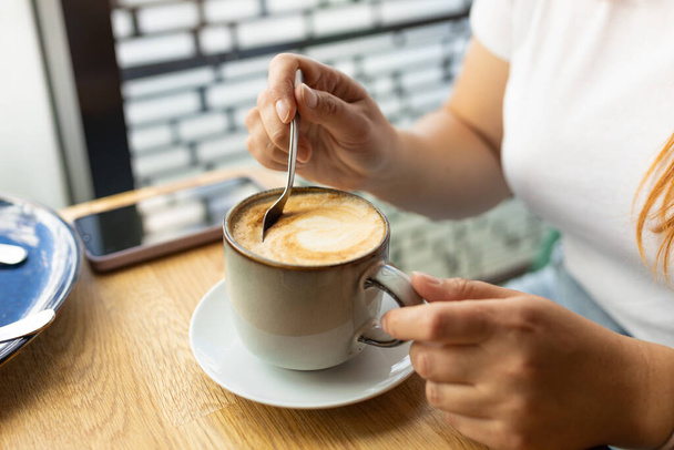 Tasse Kaffee mit Löffel und Hand auf Café-Hintergrund. Eine junge Frau trinkt Kaffee und sitzt tagsüber an einem Holztisch im Café. Frauenhände rühren mit Löffel Cappuccino-Schaum - Foto, Bild