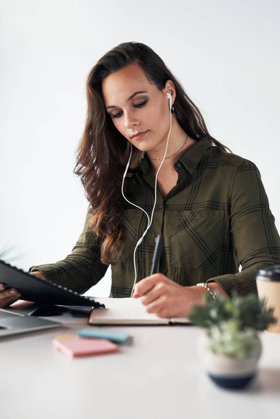 Music helps me work better. an attractive young businesswoman listening to music through earphones while making notes in a modern office - Photo, Image