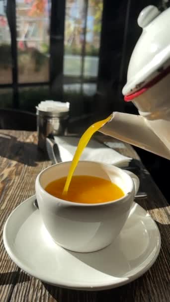 Hot vitamin sea buckthorn tea with steam pours by stream from teapot in teacup with saucer standing on old wooden table. Concept of natural hot drinks during seasonal flu and cold. Slow motion. - Footage, Video