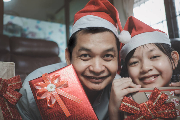 Ασιατική οικογένεια αποτελούμενη από πατέρα και κόρη φορούν καπέλα Αϊ-Βασίλη και δίνουν δώρα ο ένας στον άλλο κατά τη διάρκεια του φεστιβάλ. "Χριστούγεννα" και Ευτυχισμένο το νέο έτος στο σπίτι - Φωτογραφία, εικόνα