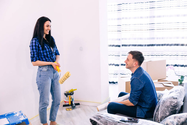 młoda pozytywna para zabawiająca się podczas remontu i malowania ścian w mieszkaniu. Mężczyzna siedzi na kanapie i dziewczyna z wałkiem do malowania w ręku - Zdjęcie, obraz
