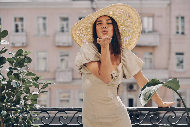 Full length of beautiful young woman in elegant hat blowing a kiss while relaxing on the balcony - Фото, зображення