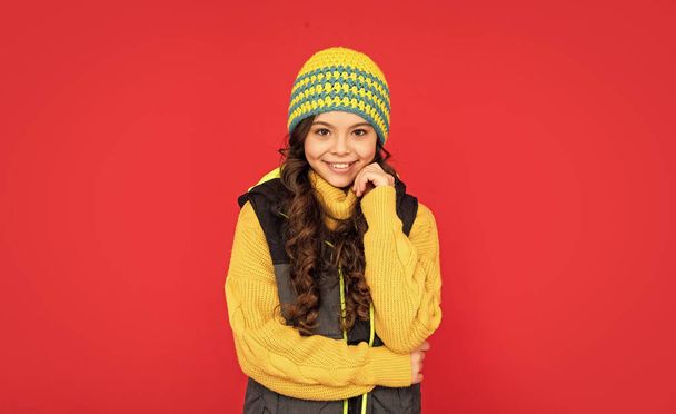 portrait of child wearing warm clothes. express positive emotion. winter fashion. happy kid with curly hair in hat. teen girl on red background. - Photo, image