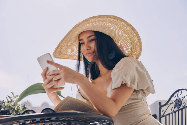 Beautiful young woman in elegant hat using smart phone while relaxing on the balcony - Photo, Image