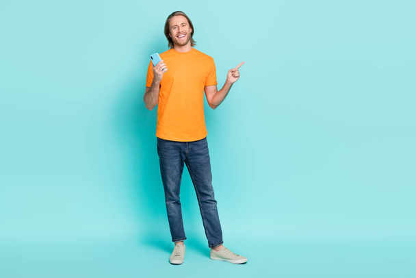 Full length photo of positive funny man wear orange t-shirt walking writing modern gadget empty space isolated turquoise color background. - Photo, Image