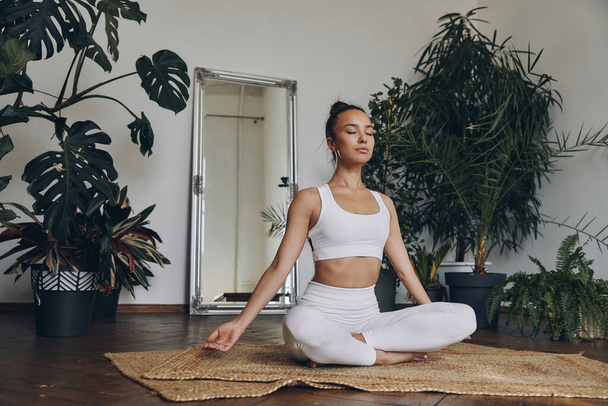 Beautiful young woman meditating while sitting on the floor with houseplants all around her - Foto, Bild