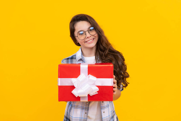 cheerful teen girl in glasses with present. studio shot of teen girl hold present. teen girl holding box of present. teen girl with present isolated on yellow background. - Photo, Image