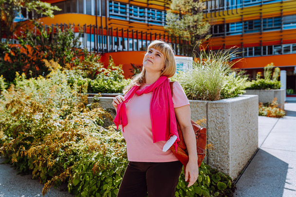 жінка середнього віку, світле довге волосся, в яскраво-рожевому шарфі, літня футболка, вивчає нову будівлю з цікавістю і оціночним виглядом, на тлі архітектури. житлова нерухомість, купити оренду
 - Фото, зображення