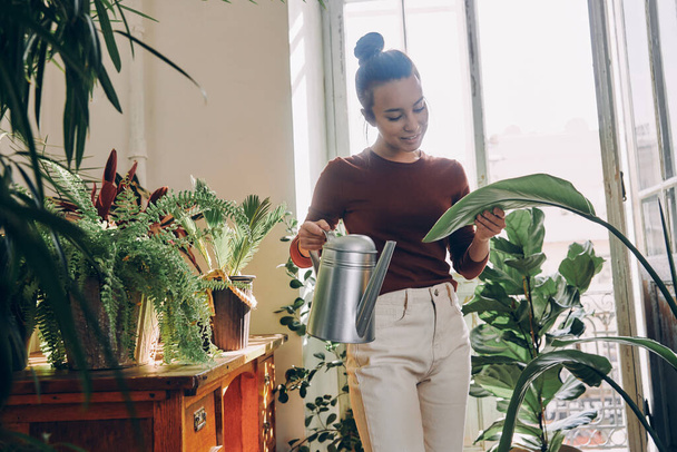 Attractive young woman watering houseplants and smiling while standing at the domestic room - Photo, image