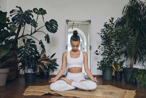 Concentrated young woman meditating while sitting on the floor with houseplants all around her - Photo, Image