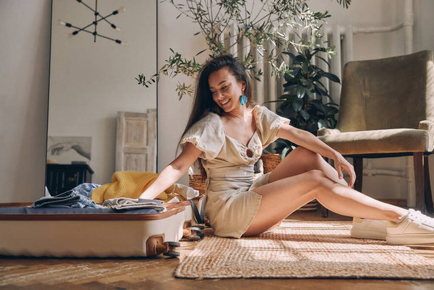 Elegant young woman packing clothes to suitcase and smiling while sitting on the floor at home - Foto, Imagen