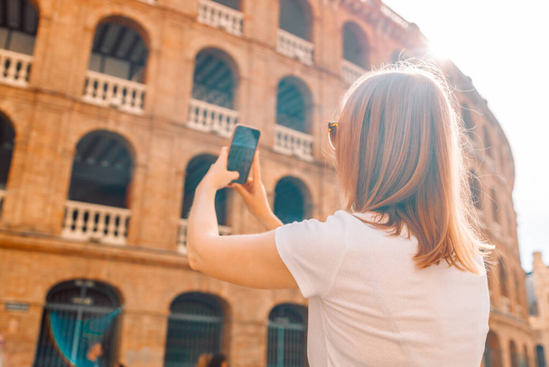 Vista trasera de una turista rubia tomando fotos en su teléfono móvil mientras camina por el casco antiguo de Plaza de Toros de Valencia. Vista desde atrás.  - Foto, Imagen