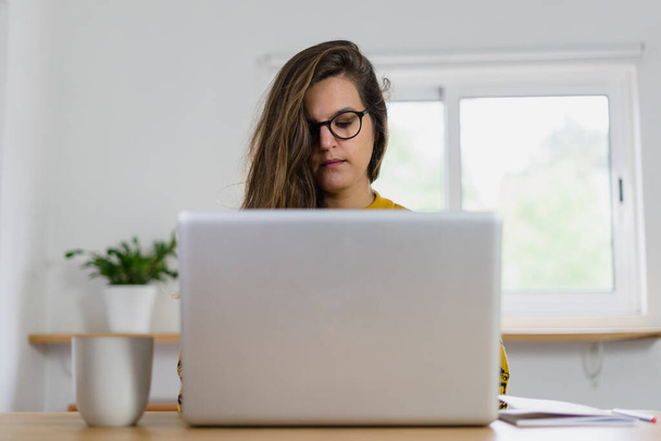 Woman with glasses working on her laptop seen from the front. White mug on your right and notebook on your left. High quality photo - Фото, изображение