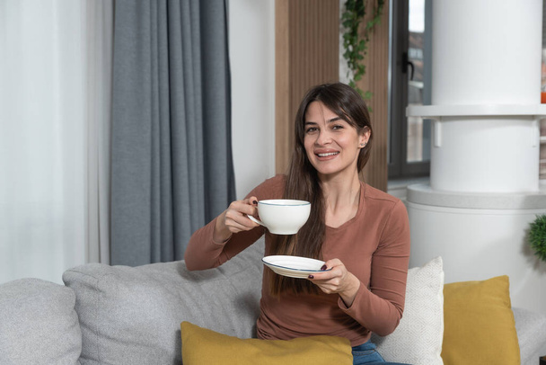 Young satisfied smiling woman drinking coffee or tea in her cozy new apartment. Female starting new life at new home sitting on sofa looking at camera. - Foto, imagen