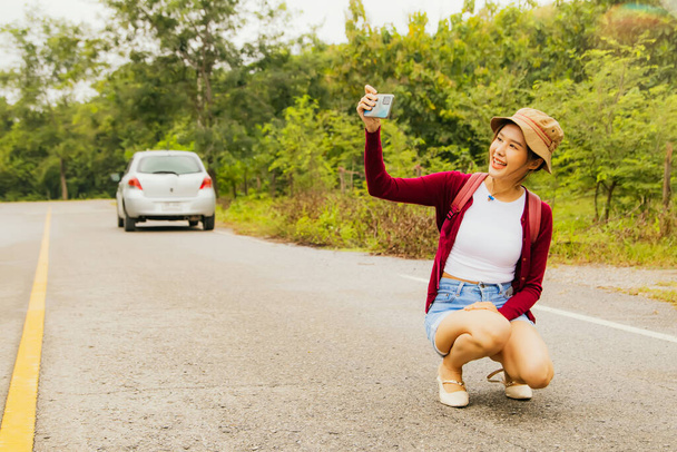 Retrato bonito menina sorrindo bonito olhando para selfie câmera smartphone com férias felizes: Mulher asiática vestida com uma roupa privada está feliz em sentar-se e tirar uma selfie em uma estrada sem carros em execução. - Foto, Imagem