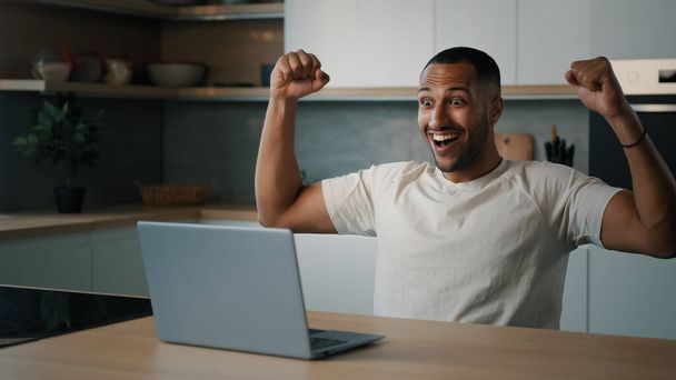 African 30s facet mężczyzna siedzi przy stole w kuchni szuka w laptopie gry online wygrać uczucie zwycięzca cieszyć się zwycięstwem otrzymać nagrodę wzrost finansowy w zakładzie świętować zakłady podekscytowany dobrą wiadomością - Zdjęcie, obraz