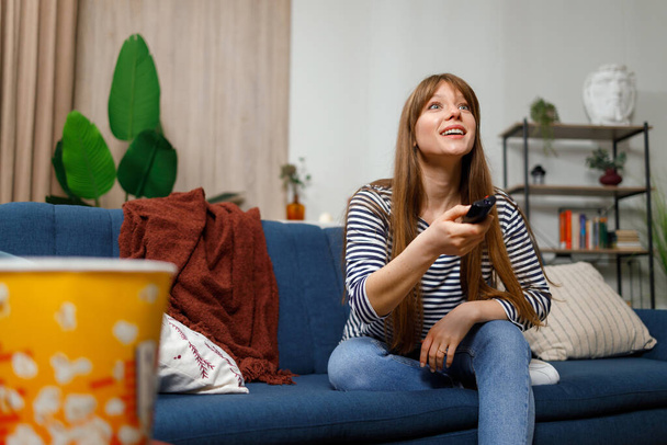Χαρούμενη γυναίκα που κάθεται στον καναπέ αλλάζει κανάλια στην τηλεόραση αναζητώντας ενδιαφέροντα προγράμματα. Εθισμός στην τηλεόραση - Φωτογραφία, εικόνα