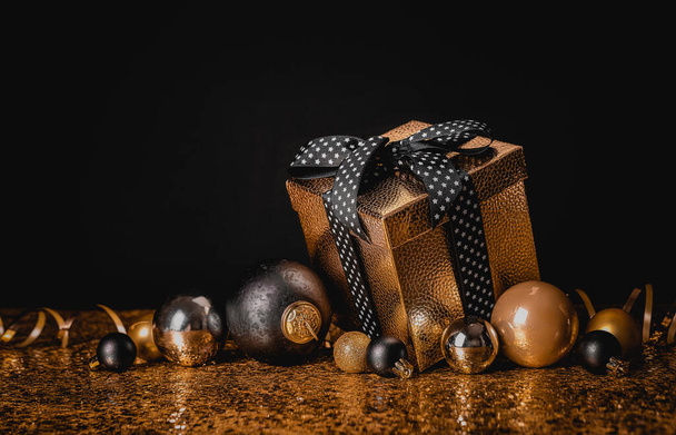 Congratulation Merry Christmas and Happy New Year. Golden box with a gift and Christmas balls on a black and gold background - Photo, Image