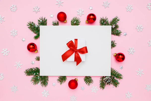 Рождество, новогодние праздники. Белая подарочная коробка с красным бантом в рамке из натуральных елок и украшения на белом и пастельно-розовом фоне - Фото, изображение