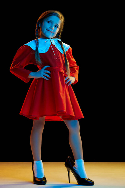 Fashion show. Portrait of beautiful cute little girl with pigtails wearing red festive dress and heels isolated over dark background. Concept of children emotions, child psychology - Valokuva, kuva
