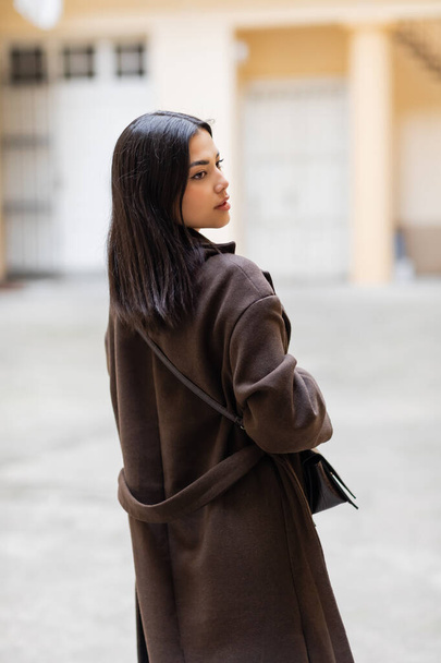 back view of young brunette woman in trendy coat with crossbody looking away outdoors in prague - Φωτογραφία, εικόνα