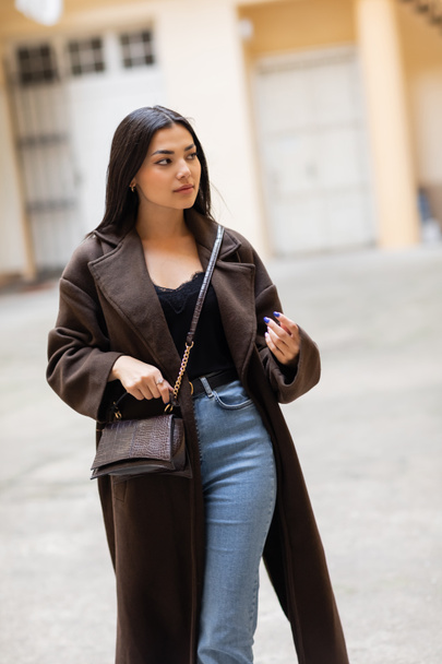 young woman in coat and jeans holding crossbody on city street in prague - 写真・画像