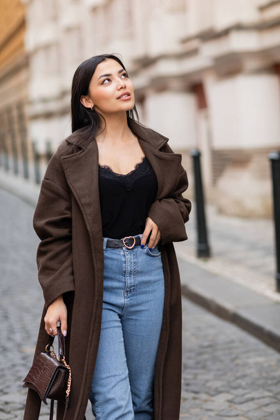 young and pretty woman in trendy coat holding handbag and looking away in prague on urban street - Zdjęcie, obraz