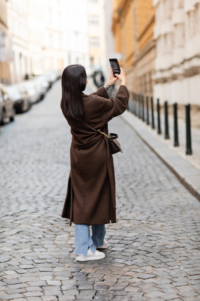 back view of brunette woman in brown coat taking photo on street in prague - Photo, image