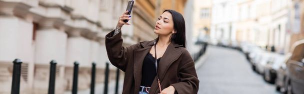 pretty brunette woman taking photo on cellphone on blurred street in prague, banner - Foto, Imagen