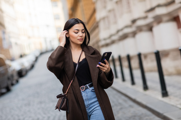 young woman in brown coat fixing hair and looking at cellphone on blurred street in prague - Foto, afbeelding