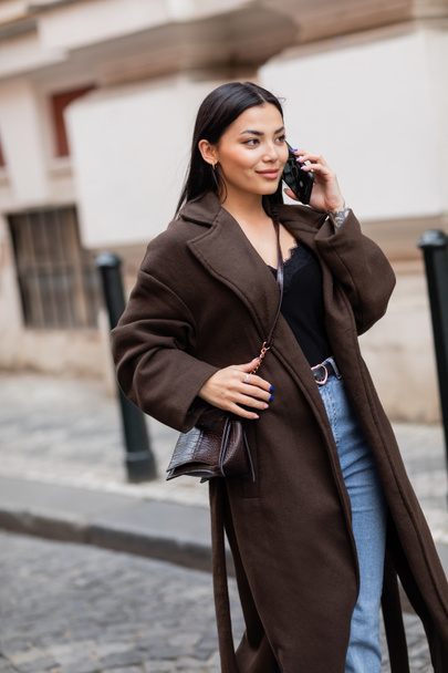 Lächelnde brünette Frau im schicken Mantel, die auf der verschwommenen Straße in Prag mit dem Smartphone spricht - Foto, Bild