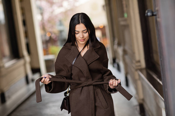 young brunette woman tying coat belt on urban street in prague - Foto, immagini