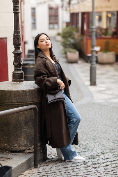 full length of brunette woman in trendy coat with crossbody looking away near lamppost in prague - Zdjęcie, obraz