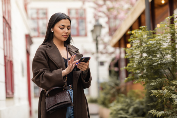 brunette woman in coat and wireless earphone using smartphone near green plants on street in prague - Photo, Image