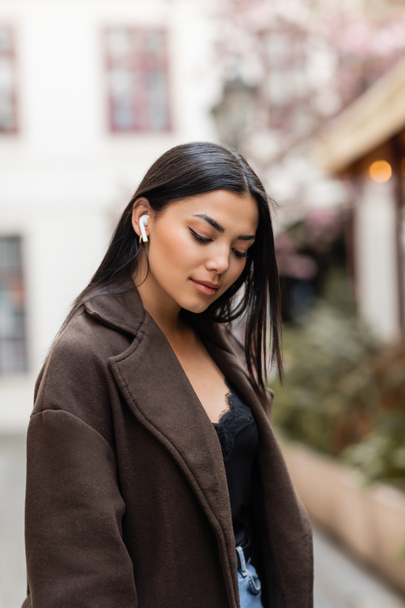 young brunette woman in stylish coat listening music in wireless earphone on street in prague - 写真・画像