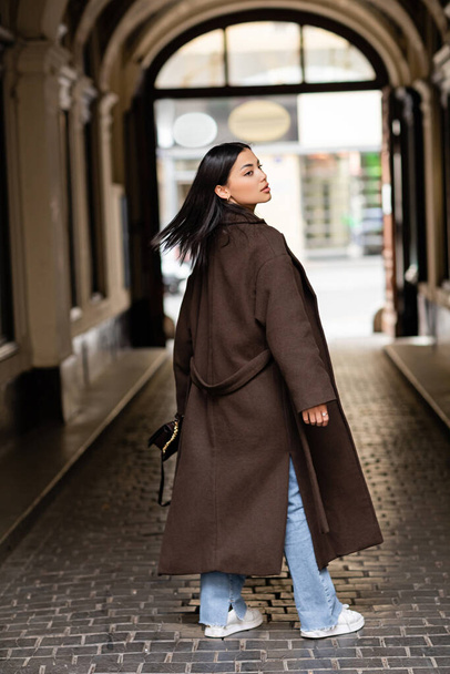 back view of young brunette woman in brown coat looking away under arch building in prague - Foto, Imagem