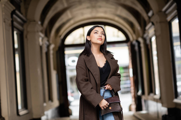 brunette woman in coat holding handbag and looking away near arch building in prague - Фото, изображение
