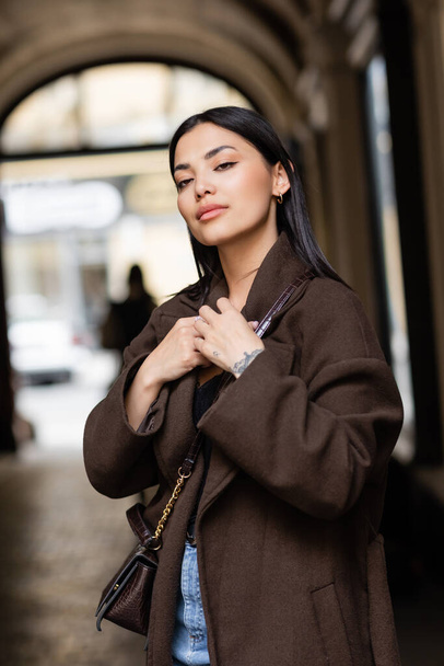 young brunette woman in trendy brown coat with crossbody on blurred background in prague - Φωτογραφία, εικόνα