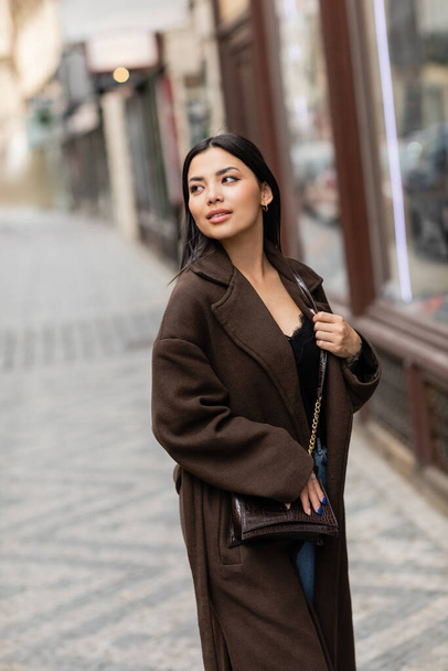 pretty brunette woman in autumn coat holding crossbody and looking away on blurred street in prague - Photo, Image